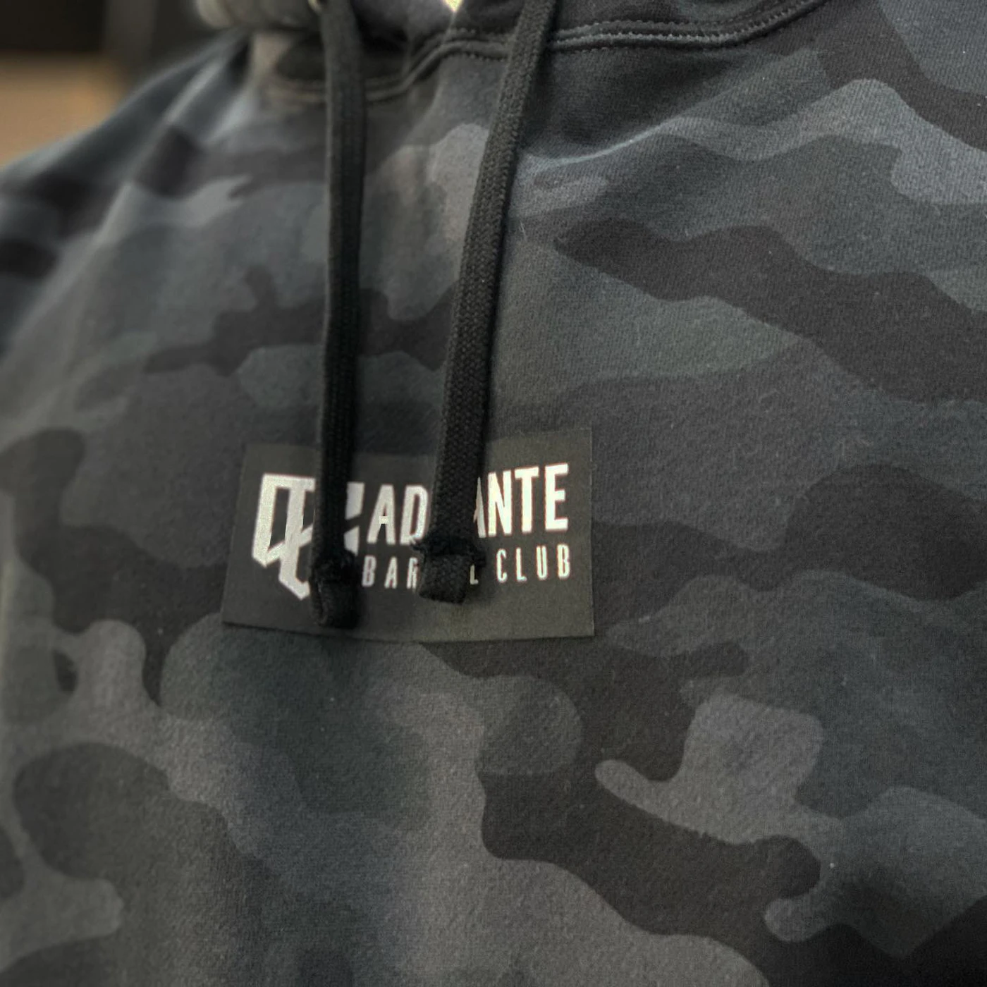 Front hoodie view of Adelante Barbell Club's shield logomark and wordmark printed on a dark gray camo hoodie
