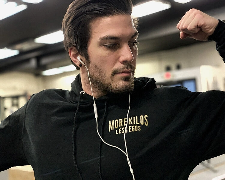 Man flexing and wearing an Adelante Barbell Club 2019 Hoodie, front pocket design says "More Kilos, Less Egos"